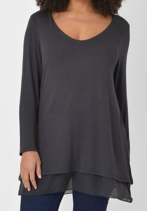 Vrouw draagt een donkergrijze gelaagde tuniek met lange mouwen, een ronde hals en blauwe spijkerbroek.