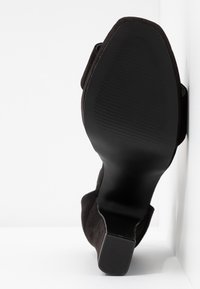 Chaussure à talons hauts en velours noir avec une semelle en caoutchouc lisse et plate, et une texture nervurée subtile près du talon. Détail de sangle visible sur le côté.