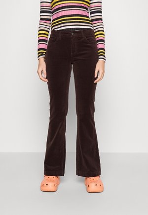 Person iført mørkebrune fløjlsbukser, en langærmet top med flerfarvede striber og orange slip-on sko, stående mod en ensfarvet baggrund.