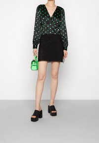 Jupe portefeuille noire associée à un chemisier vert à pois avec manches longues. Le mannequin porte des mules noires à talons épais et tient un sac à main vert.