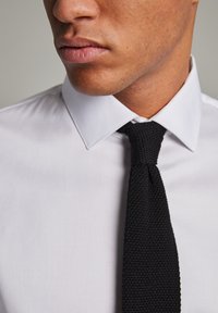 Camisa blanca de vestir con cuello clásico, contrastada por una corbata de punto negra texturizada. Vista en primer plano del área del cuello y la corbata.