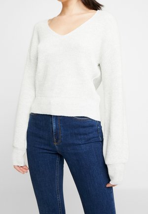 Nainen pukeutuneena valkoiseen pitkähihainen V-kaulaiseen neulepaitaan, yhdistettynä tummansinisiin farkkuihin, yksinkertaisen taustan edessä.