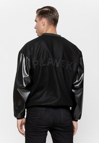 Czarna kurtka bomberka z czarnymi skórzanymi rękawami i teksturowanym materiałem, z haftem "PILAWSKI" na plecach.
