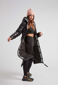 Mujer con un sujetador deportivo negro, mallas, zapatillas, un gorro rosa y una larga chaqueta de plumas negra abierta con la marca "JACKIT" por dentro.