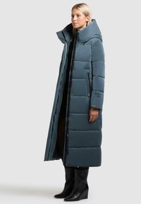 Lang pufferjakke i dæmpet teal, med høj krave, chunky quiltedesign og lynlåslommer i siden, parret med sorte ankelsko.