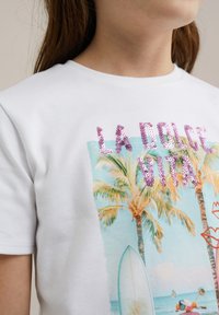 Bela bomba iz bombažna majica z večbarvno grafiko palm, okrašena s sekvencami napisa "LA DOLCE VITA" v roza barvi, z ilustracijo surf deske.