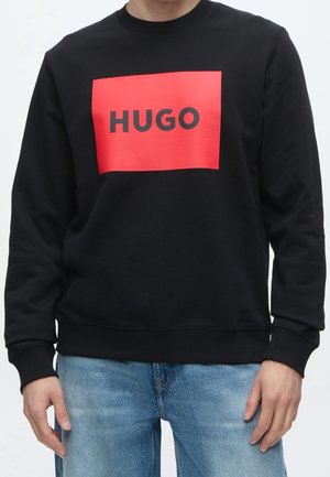 Osoba nosząca czarną bluzę z czerwonym prostokątnym logo HUGO i jasnoniebieskie dżinsy, stojąca z rozluźnionymi rękami.