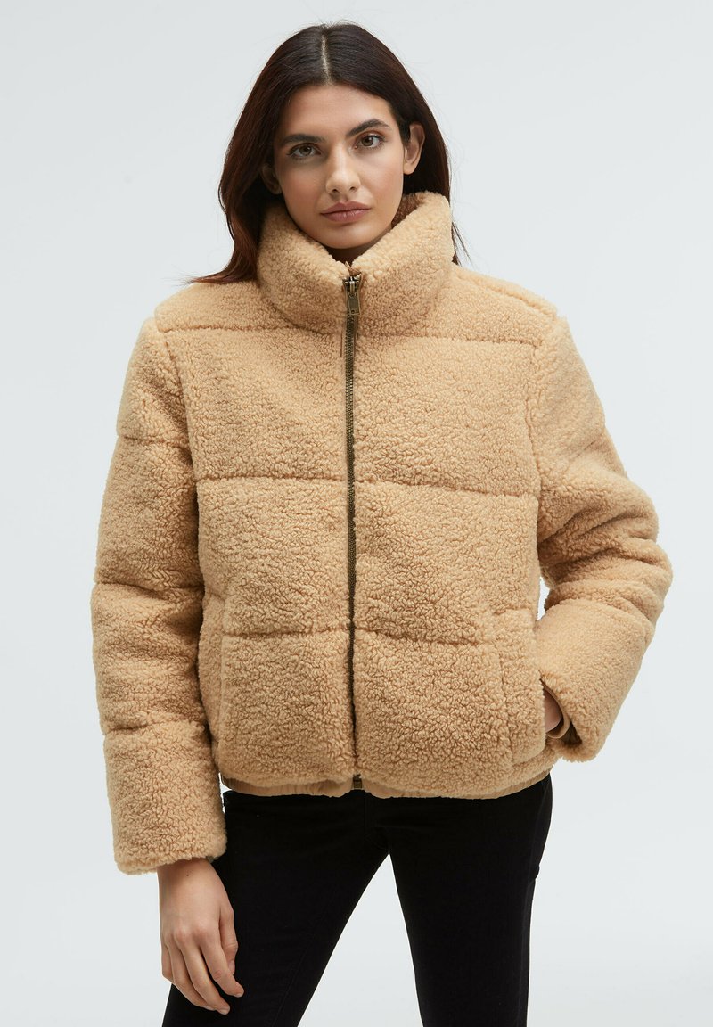 Beige faux fur jacket with a high collar, featuring a front zipper, boxy design, and horizontal quilted pattern. Soft texture and relaxed fit.