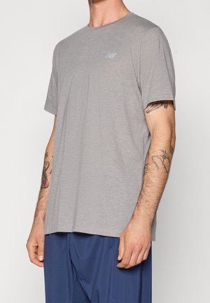 Mężczyzna ubrany w prostą szarą koszulkę na krótki rękaw marki New Balance i niebieskie spodnie, pokazujący tatuaże na obu przedramionach na białym tle.