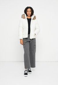 Vrouw in een witte gewatteerde jas met een met bont gevoerde capuchon, zwarte top, grijze wijde cargobroek en zwart-witte sneakers, staand tegen een witte achtergrond.