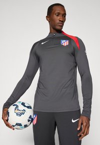 Szürke, hosszúujjú sportpulóver piros vállsávokkal és cipzáros nyakkal, klublogóval. A modell egy mintás focilabdát tart.