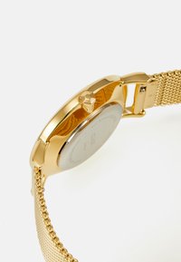 Montre en acier inoxydable doré avec un bracelet en mailles, un boîtier rond et une couronne texturée avec une surface lisse et réfléchissante à l'arrière.