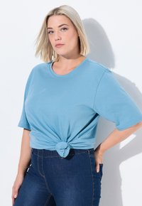 Una maglietta azzurro chiaro a maniche corte presenta un design con nodo sulla parte anteriore, realizzata in tessuto morbido, abbinata a jeans in denim scuro.