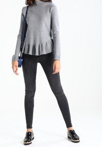 Maglietta grigia a maniche lunghe con orlo a peplo, abbinata a jeans skinny neri e scarpe nere lucide. Una borsa a tracolla blu è indossata sulla spalla sinistra.