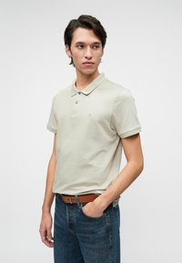 Világosszürke pamut póló, gallérral, két gombbal és logóval. Kék farmernadrággal és barna övvel párosítva.