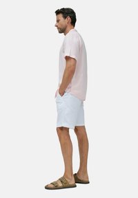 Lichtroze shirt met korte mouwen, witte shorts en bruine sandalen. Het shirt heeft een ontspannen pasvorm met een subtiele textuur en zijsplitsingen.