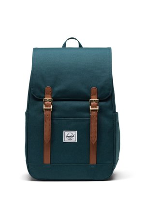 Mochila de tela color verde azulado con correas de cuero marrón y herrajes dorados. Cuenta con un bolsillo frontal y un parche con el logotipo. Diseño rectangular y robusto.