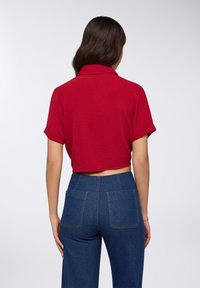 Polo shirt rossa corta con una finitura testurizzata, caratterizzata da maniche corte arrotolate. Abbinata a pantaloni in denim blu a vita alta con tasche posteriori.
