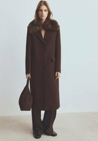 Bruin wollen jas met een nepbonten kraag, voorzien van een getailleerd ontwerp, voorzakken en zes knopen, gecombineerd met donkerbruine broek en tas.