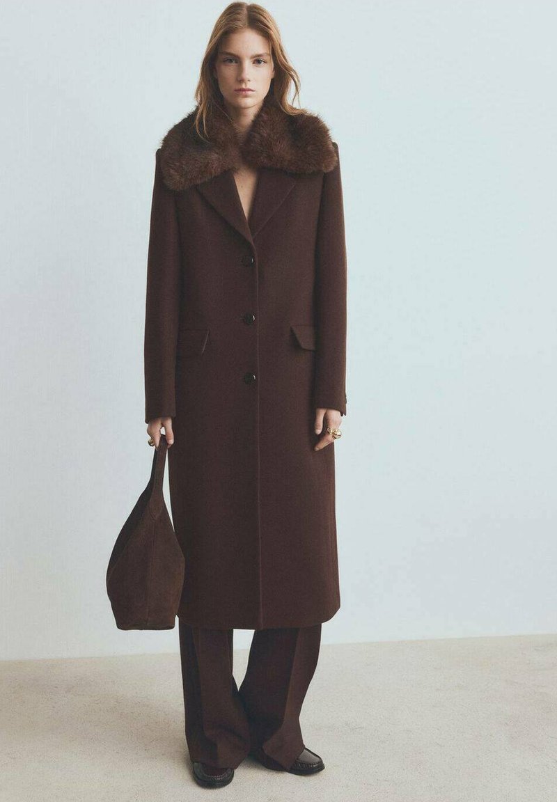 Bruin wollen jas met een nepbonten kraag, voorzien van een getailleerd ontwerp, voorzakken en zes knopen, gecombineerd met donkerbruine broek en tas.