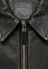 Black leather jacket featuring a pointed collar, central zipper, and a branded tag with "MNG LEATHER" embossed on it. Textured surface detail visible.