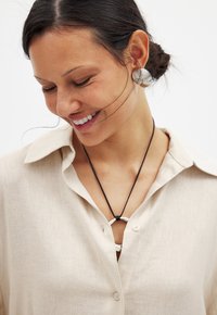 Camicia button-up in lino beige con colletto, indossata con una collana in corda nera che presenta un accento circolare in argento. Orecchini in argento texturizzato.