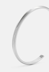 Sølv metal armbånd med en glat overflade og teksturerede diagonale linjer, der har en afrundet åben ende og et minimalistisk design.