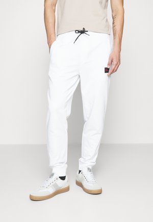Pantalones de chándal blancos con bolsillos laterales, puños elásticos y una cinturilla con cordón. Presenta un pequeño parche de logo rojo y negro en la cadera.