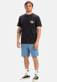 Svart bomulls t-skjorte med et fargerikt grafisk motiv på brystlommen, kombinert med lyseblå shorts og svarte joggesko med hvite detaljer.
