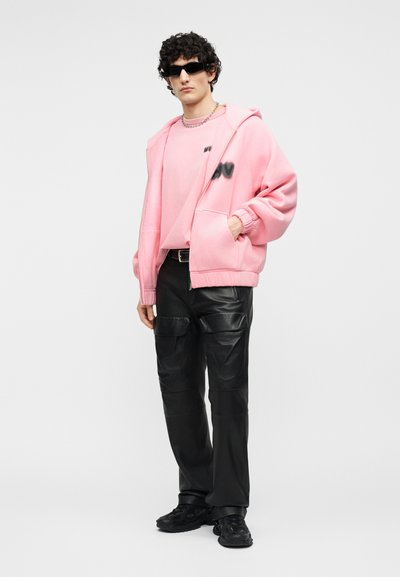 Homme portant des lunettes de soleil noires, un sweat à capuche rose surdimensionné et un t-shirt, un pantalon cargo en cuir noir et des baskets noires, debout les mains dans les poches.