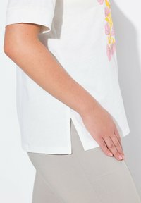 T-shirt en coton blanc avec un design floral rose et jaune, à manches courtes, fentes sur les côtés, porté avec un pantalon ajusté gris clair.