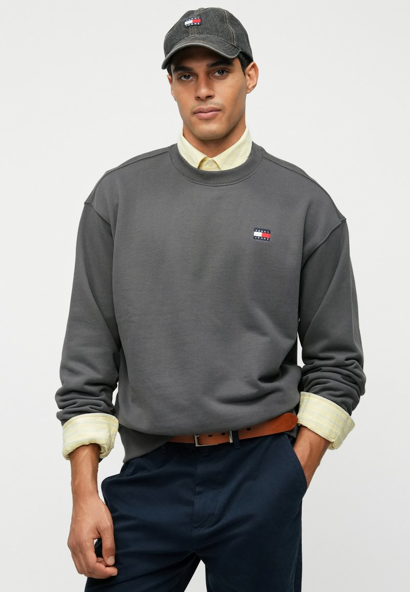 Dark grey sweatshirt with ribbed neckline, featuring a small logo patch. Layered over a light plaid shirt with rolled cuffs, paired with navy pants.