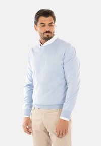 Maglione lavorato a maglia azzurro chiaro con scollo rotondo, maniche lunghe e una texture morbida, abbinato a pantaloni beige e una camicia bianca con colletto sotto.