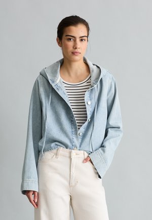 Jonge vrouw draagt een lichtblauwe spijkerjas met capuchon over een zwart-wit gestreept shirt en crèmekleurige broek tegen een effen achtergrond.