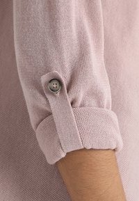 Hellrosa gestrickter Pulloverärmel mit einem detailreichen, geknöpften Bündchen. Weiche Textur und lässige Passform, zeigt Rippstrick und glatte Oberfläche.