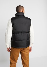 Man i svart vadderad väst, vit långärmad skjorta och bruna byxor, stående vänd bort från kameran mot en neutral bakgrund.