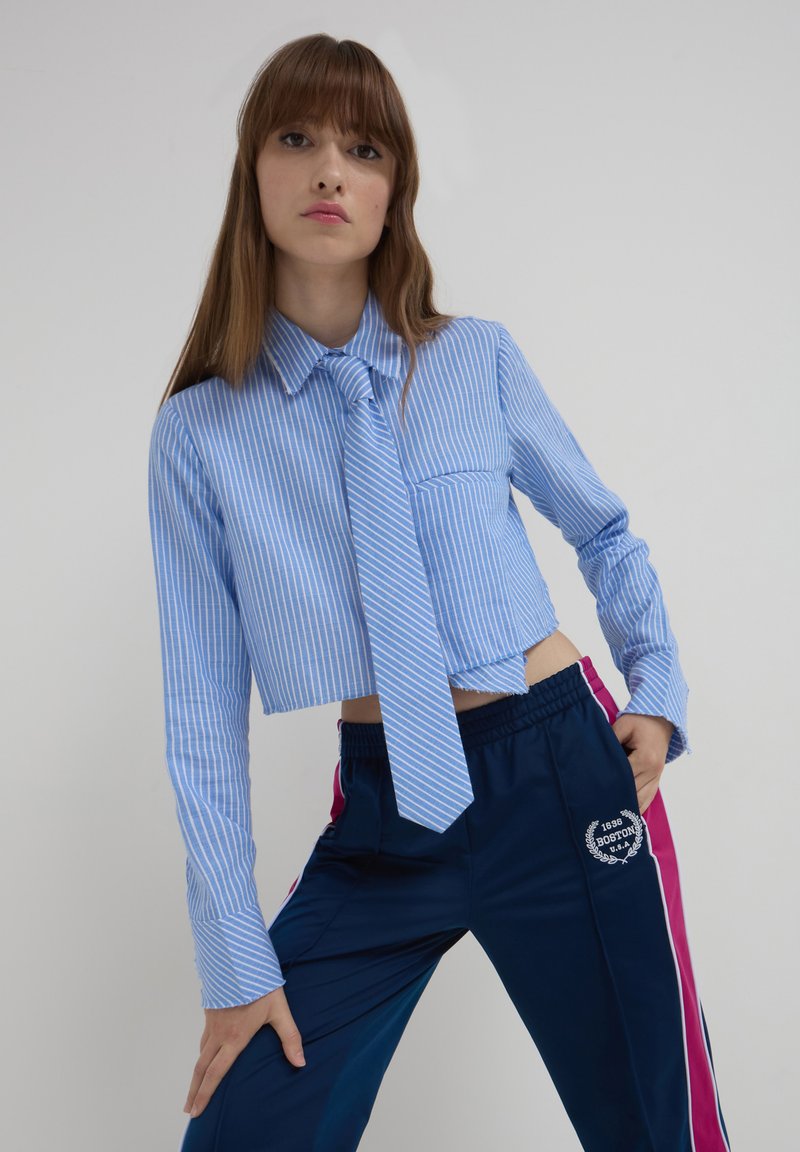 Junge Frau in einem blau gestreiften, kurz geschnittenen Hemd und einer Krawatte, navyblauen Sporthosen mit rosa Seitenstreifen, die vor einem einfarbigen Hintergrund posiert.