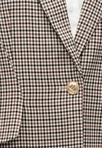 Blazer à motif pied-de-poule marron et blanc avec des accents noirs, comprenant un bouton en or et une poche plaquée sur le côté gauche.