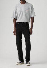 Homme portant un t-shirt gris clair à manches courtes avec poche et étiquette, pantalon noir et baskets noires avec semelles blanches et lacets blancs.