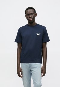 Μπλε βαμβακερό μπλουζάκι με κοντά μανίκια και στρογγυλή λαιμόκοψη, με μικρή λεπτομέρεια λογότυπου στο αριστερό στήθος. Φοριέται με ανοιχτόχρωμα μπλε τζιν.