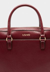 Bordeaux læder håndtaske med tekstureret overflade, guld hardware detaljer og tophåndtag. Har logo detaljer og lynlåslukning.