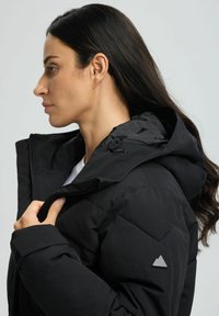 Frau im Profil, die den Kragen einer schwarzen Winterjacke mit Kapuze und gesteppten Ärmeln mit triangle Logo am Arm anpasst.