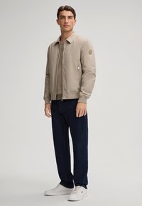 Beżowa kurtka bombowa z zamkiem z przodu, ściągaczami przy mankietach i kieszeniami bocznymi. Nosi się ją na brązowym swetrze, w połączeniu z granatowymi dżinsami i białymi trampkami.