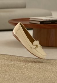 Roomkleurige leren loafers met een gestikt ontwerp, gouden accenten, een rubberen zool en een gladde textuur, zwevend boven een lichte achtergrond.