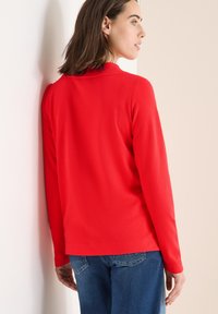 Röd långärmad topp med avslappnad passform, rundad halsringning och slät textur, i kombination med blå denimjeans. Bakifrån mot en ljus bakgrund.
