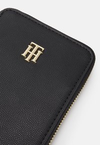 Musta, teksturoitu lompakko, jossa on kultainen leima-logo ja kultainen vetoketjutekstu reunassa. Yksinkertainen ja tyylikäs muotoilu.