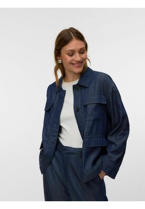 Kvinde smiler, iført en mørkeblå denimjakke og bukser med en hvid top, hænderne i lommerne, mod en ensartet lys baggrund.