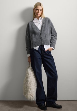 Cardigan en laine gris avec deux poches, superposé sur une chemise blanche. Associé à un jean évasé bleu foncé et un sac texturé blanc. Mocassins noirs.