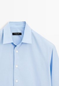 Chemise à rayures bleu clair avec un col classique, boutons blancs et une étiquette de marque noire à l'intérieur. Matière en coton avec une texture lisse.