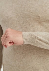 Pull beige avec des poignets côtelés, affichant un motif de tricot texturé. Gros plan sur une main reposant contre le tissu.